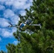 A multi-rotor drone is captured mid-flight against a partly cloudy blue sky, with pine trees visible in the background and foreground. The drone appears to be a custom or specialized hexacopter-style UAV with a white frame, featuring a blue cylindrical payload or component mounted at its center. The image is taken from a low angle looking upward, providing a clear view of the drone's underside and rotor configuration.