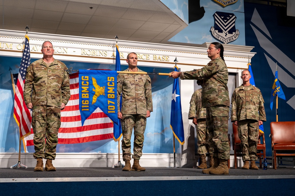 A military ceremony is taking place on a stage at Robins Air Force Base, likely a change of command or activation/inactivation ceremony. Four personnel in OCP (Operational Camouflage Pattern) uniforms are on stage, with one individual passing a unit guidon/streamer to another. The ceremony banner visible reads '116 BMCG' and '107 BMCS', indicating units associated with Bomb Maintenance Control.