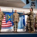A military ceremony is taking place on a stage at Robins Air Force Base, likely a change of command or activation/inactivation ceremony. Four personnel in OCP (Operational Camouflage Pattern) uniforms are on stage, with one individual passing a unit guidon/streamer to another. The ceremony banner visible reads '116 BMCG' and '107 BMCS', indicating units associated with Bomb Maintenance Control.