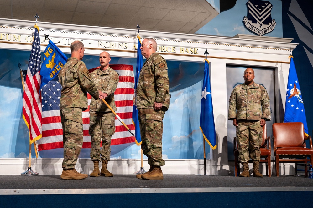 A military ceremony is taking place on a stage at the Museum of Aviation at Robins Air Force Base. Four personnel in OCP (Operational Camouflage Pattern) uniforms are on stage, with two individuals appearing to exchange a handshake, likely during a change of command or award ceremony. A unit guidon and multiple flags, including the U.S. flag and Air Force flags, are prominently displayed.