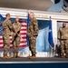 A military ceremony is taking place on a stage at the Museum of Aviation at Robins Air Force Base. Four personnel in OCP (Operational Camouflage Pattern) uniforms are on stage, with two individuals appearing to exchange a handshake, likely during a change of command or award ceremony. A unit guidon and multiple flags, including the U.S. flag and Air Force flags, are prominently displayed.