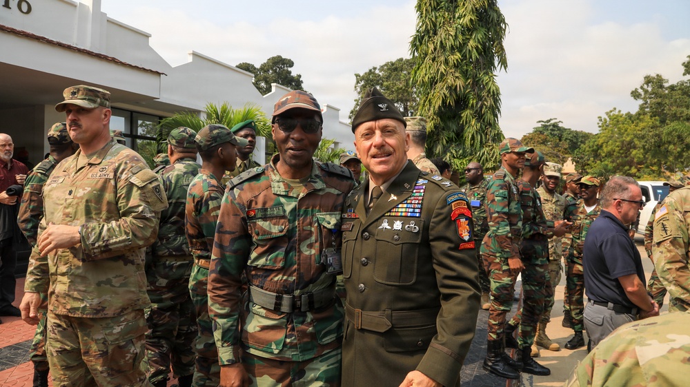 A group of military personnel from what appears to be two different nations are gathered outdoors near a white building. Two senior officers — one in a green dress uniform with numerous decorations and a garrison cap, and one in camouflage — are posing together in the foreground, suggesting an official bilateral military meeting or visit. Additional soldiers in both U.S. Army combat uniforms and foreign camouflage uniforms are visible in the background.