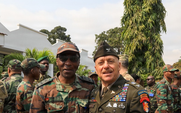 A group of military personnel from what appears to be two different nations are gathered outdoors near a white building. Two senior officers — one in a green dress uniform with numerous decorations and a garrison cap, and one in camouflage — are posing together in the foreground, suggesting an official bilateral military meeting or visit. Additional soldiers in both U.S. Army combat uniforms and foreign camouflage uniforms are visible in the background.