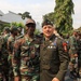A group of military personnel from what appears to be two different nations are gathered outdoors near a white building. Two senior officers — one in a green dress uniform with numerous decorations and a garrison cap, and one in camouflage — are posing together in the foreground, suggesting an official bilateral military meeting or visit. Additional soldiers in both U.S. Army combat uniforms and foreign camouflage uniforms are visible in the background.