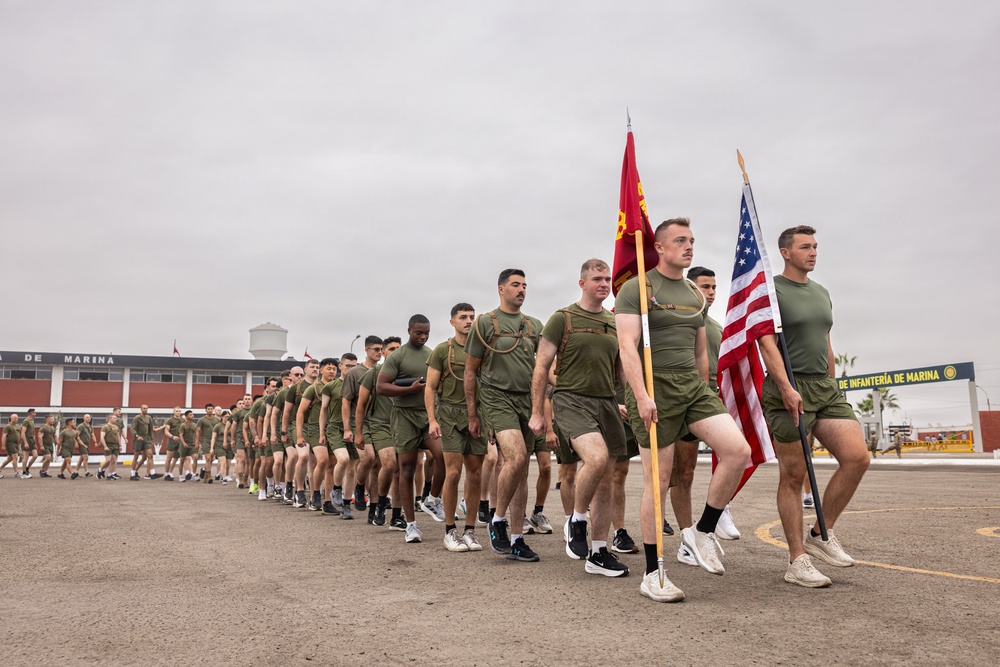 A formation of U.S. Marines in olive drab physical training (PT) uniforms marches in a column on a paved parade ground. Two flag bearers at the front carry the American flag and what appears to be a Marine Corps unit guidon. The backdrop includes a building with signage reading 'DE INFANTERÍA DE MARINA,' indicating this is a Spanish-speaking military installation, likely in Latin America.