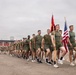 A formation of U.S. Marines in olive drab physical training (PT) uniforms marches in a column on a paved parade ground. Two flag bearers at the front carry the American flag and what appears to be a Marine Corps unit guidon. The backdrop includes a building with signage reading 'DE INFANTERÍA DE MARINA,' indicating this is a Spanish-speaking military installation, likely in Latin America.