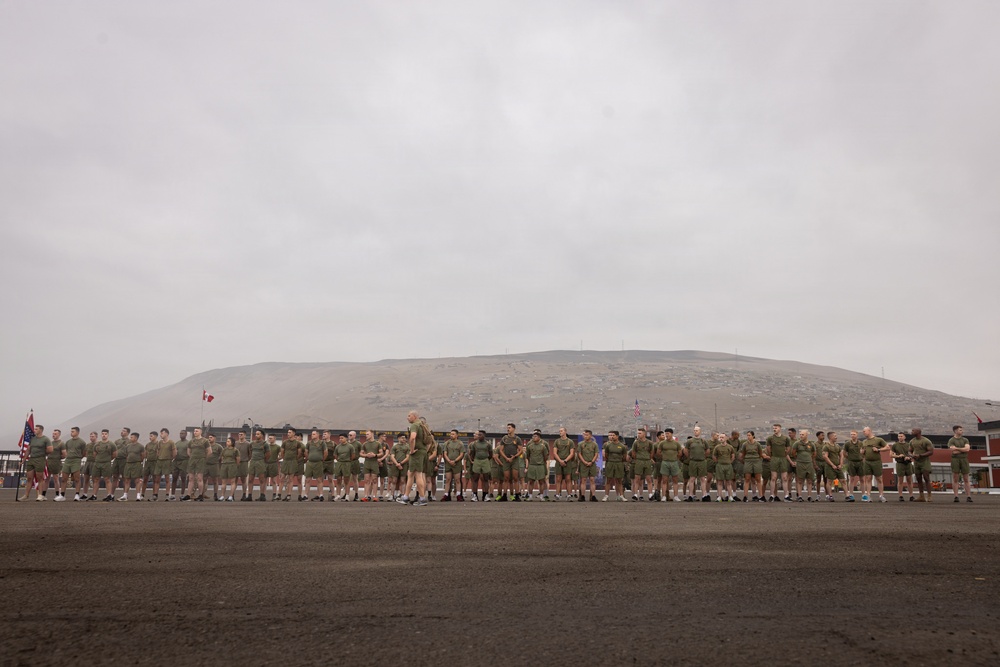 A large formation of military personnel dressed in olive green PT (physical training) uniforms are assembled on a dark gravel parade ground or tarmac. A single individual, likely an officer or NCO, stands in front of the formation appearing to address the group. The backdrop features a large barren hill or mesa under an overcast sky, with what appear to be flags including a Canadian flag and a US flag visible in the background.