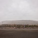 A large formation of military personnel dressed in olive green PT (physical training) uniforms are assembled on a dark gravel parade ground or tarmac. A single individual, likely an officer or NCO, stands in front of the formation appearing to address the group. The backdrop features a large barren hill or mesa under an overcast sky, with what appear to be flags including a Canadian flag and a US flag visible in the background.
