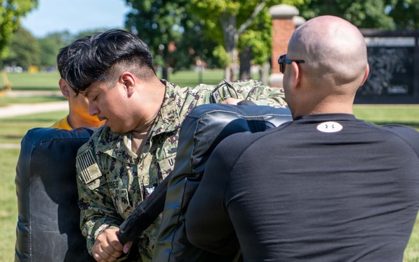 A U.S. Navy sailor in digital camouflage uniform is engaged in a physical training drill outdoors on a grassy area, practicing with a large padded training shield held by an instructor in a black Under Armour shirt. The exercise appears to be part of a military combatives or defensive tactics training session. The setting appears to be a military installation based on the brick structures and manicured grounds visible in the background.