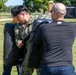 A U.S. Navy sailor in digital camouflage uniform is engaged in a physical training drill outdoors on a grassy area, practicing with a large padded training shield held by an instructor in a black Under Armour shirt. The exercise appears to be part of a military combatives or defensive tactics training session. The setting appears to be a military installation based on the brick structures and manicured grounds visible in the background.