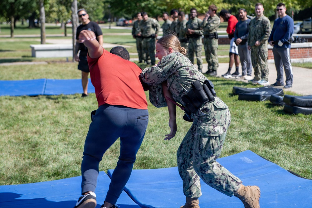 A military training exercise is taking place outdoors on blue mats, where a female service member in Navy camouflage uniform is practicing a takedown or grappling technique on a person in civilian clothes (red shirt). A group of uniformed military personnel and observers stand in the background watching the demonstration. The setting appears to be a military base or installation with green grass and trees.