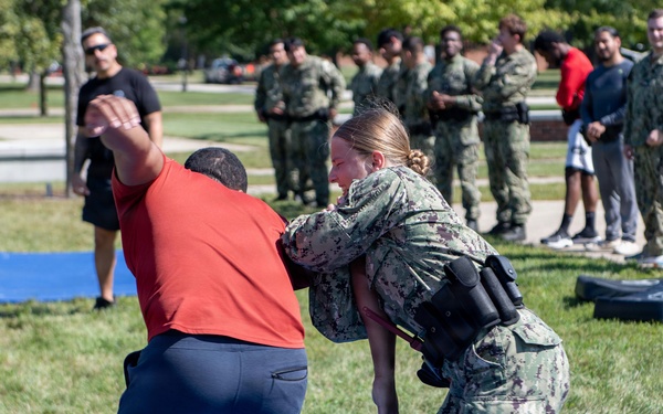 A military training exercise is taking place outdoors on blue mats, where a female service member in Navy camouflage uniform is practicing a takedown or grappling technique on a person in civilian clothes (red shirt). A group of uniformed military personnel and observers stand in the background watching the demonstration. The setting appears to be a military base or installation with green grass and trees.