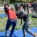 A military training exercise is taking place outdoors on blue mats, where a female service member in Navy camouflage uniform is practicing a takedown or grappling technique on a person in civilian clothes (red shirt). A group of uniformed military personnel and observers stand in the background watching the demonstration. The setting appears to be a military base or installation with green grass and trees.