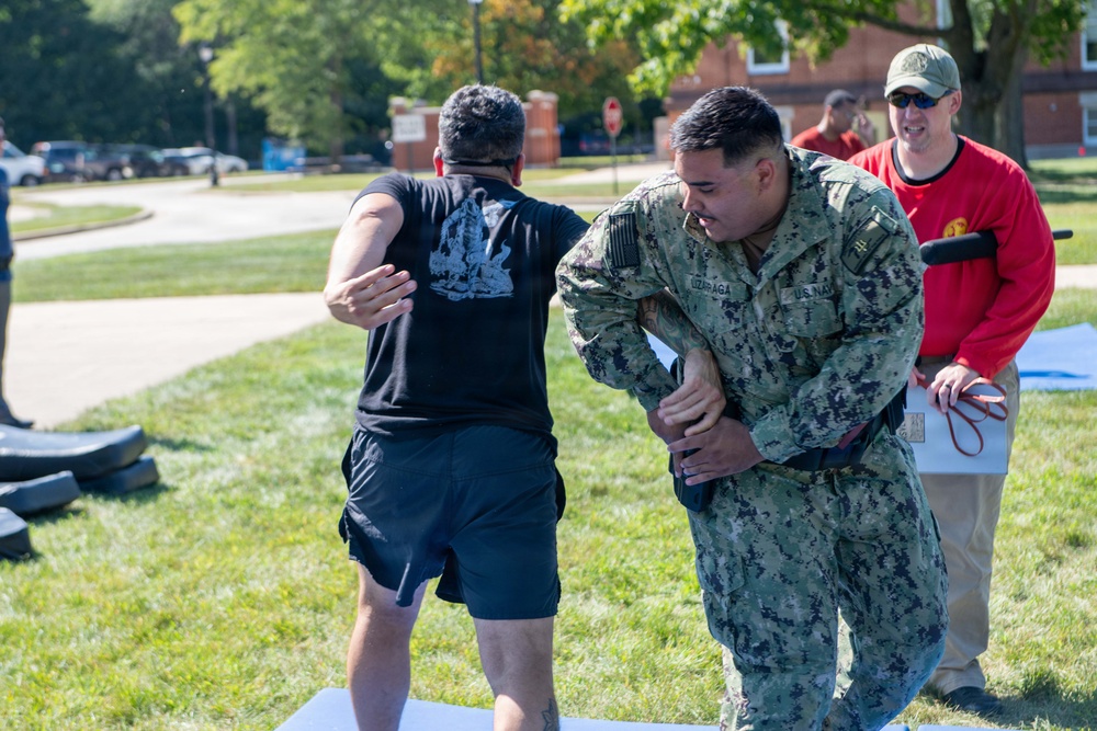 A U.S. Navy sailor in NWU (Navy Working Uniform) camouflage is performing what appears to be a combatives or self-defense training drill with a civilian or role-player dressed in black civilian clothing. A third person in a red shirt, wearing sunglasses and a cap, observes and appears to be supervising or evaluating the exercise. The setting is an outdoor grassy area on what appears to be a military installation.
