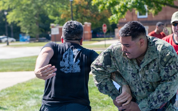 A U.S. Navy sailor in NWU (Navy Working Uniform) camouflage is performing what appears to be a combatives or self-defense training drill with a civilian or role-player dressed in black civilian clothing. A third person in a red shirt, wearing sunglasses and a cap, observes and appears to be supervising or evaluating the exercise. The setting is an outdoor grassy area on what appears to be a military installation.