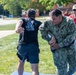 A U.S. Navy sailor in NWU (Navy Working Uniform) camouflage is performing what appears to be a combatives or self-defense training drill with a civilian or role-player dressed in black civilian clothing. A third person in a red shirt, wearing sunglasses and a cap, observes and appears to be supervising or evaluating the exercise. The setting is an outdoor grassy area on what appears to be a military installation.