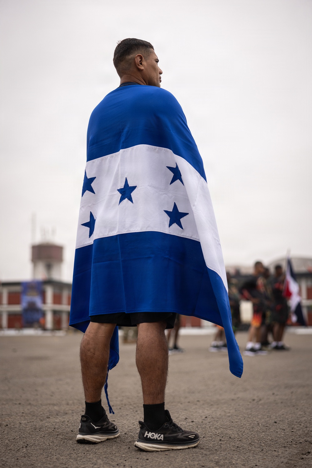 A man stands with his back to the camera, draped in the flag of Honduras — characterized by two blue horizontal stripes, a white center stripe, and five blue stars arranged in an X pattern. He is wearing black shorts, black socks, and HOKA brand running shoes. The setting appears to be an outdoor paved area with other people visible in the blurred background.