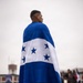 A man stands with his back to the camera, draped in the flag of Honduras — characterized by two blue horizontal stripes, a white center stripe, and five blue stars arranged in an X pattern. He is wearing black shorts, black socks, and HOKA brand running shoes. The setting appears to be an outdoor paved area with other people visible in the blurred background.