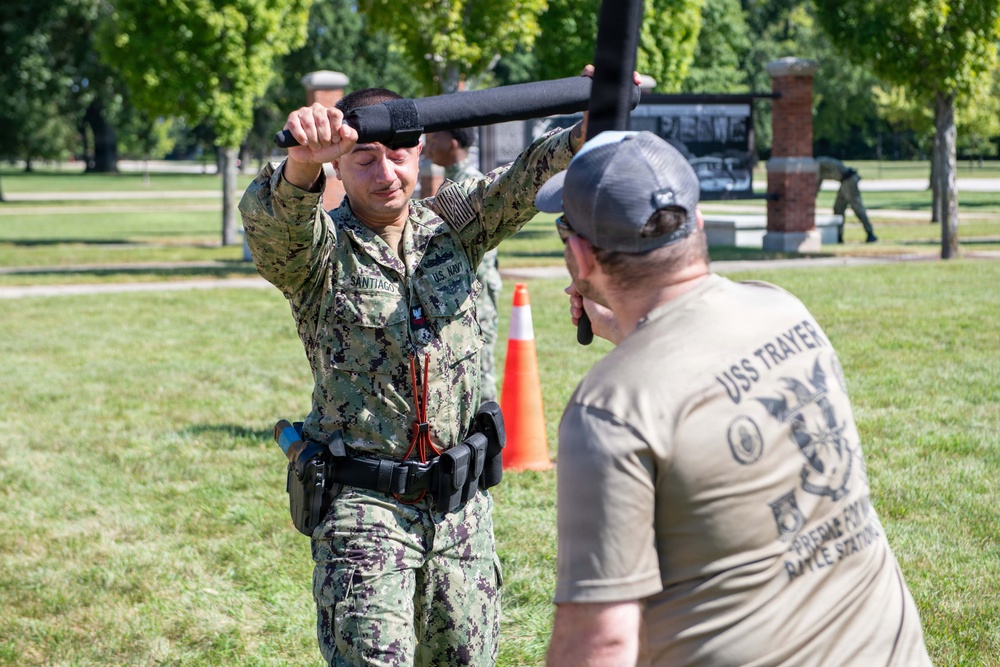 A U.S. Navy sailor in digital camouflage uniform is engaged in a baton/defensive tactics training exercise outdoors on a grassy area. The sailor, identified by the name tape 'SANTIAGO' and 'U.S. NAVY' patch, is blocking or deflecting a padded training baton held by an instructor wearing a USS Trayer t-shirt. The setting appears to be a military base training ground with other personnel visible in the background.