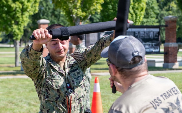 A U.S. Navy sailor in digital camouflage uniform is engaged in a baton/defensive tactics training exercise outdoors on a grassy area. The sailor, identified by the name tape 'SANTIAGO' and 'U.S. NAVY' patch, is blocking or deflecting a padded training baton held by an instructor wearing a USS Trayer t-shirt. The setting appears to be a military base training ground with other personnel visible in the background.