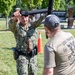 A U.S. Navy sailor in digital camouflage uniform is engaged in a baton/defensive tactics training exercise outdoors on a grassy area. The sailor, identified by the name tape 'SANTIAGO' and 'U.S. NAVY' patch, is blocking or deflecting a padded training baton held by an instructor wearing a USS Trayer t-shirt. The setting appears to be a military base training ground with other personnel visible in the background.