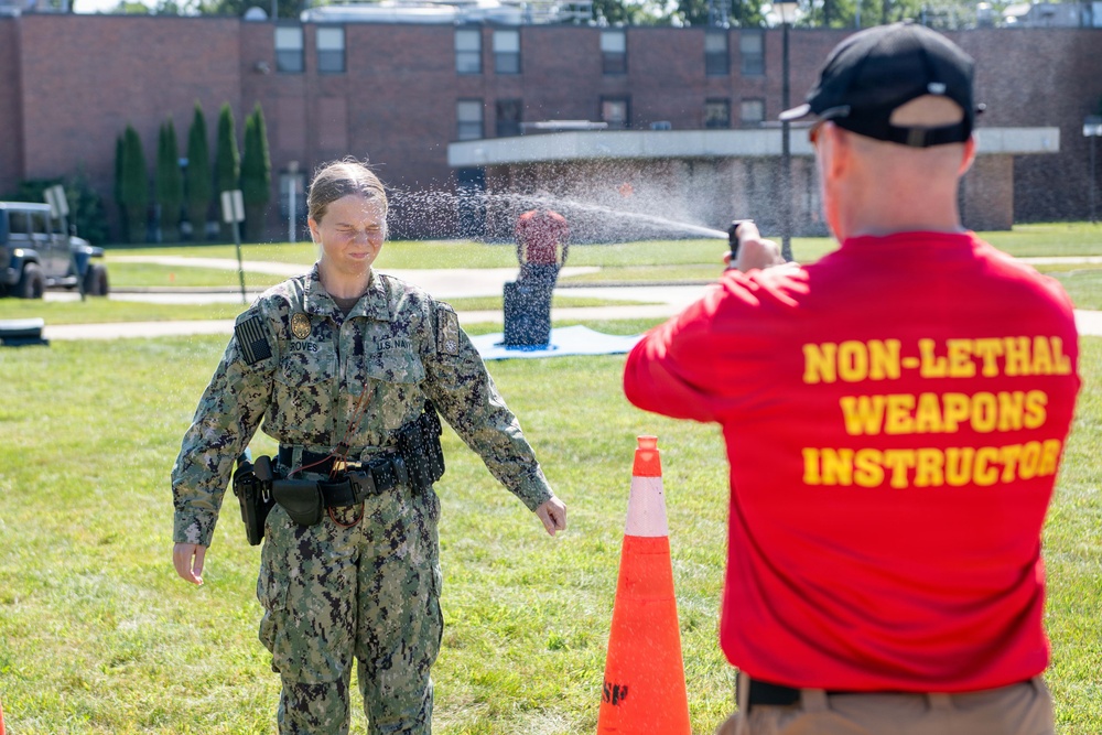 A U.S. Navy personnel member is being sprayed with what appears to be OC (pepper) spray or a similar non-lethal substance as part of a training exercise. A Non-Lethal Weapons Instructor in a red shirt is administering the spray. The training is taking place outdoors on a grassy area of what appears to be a military installation.