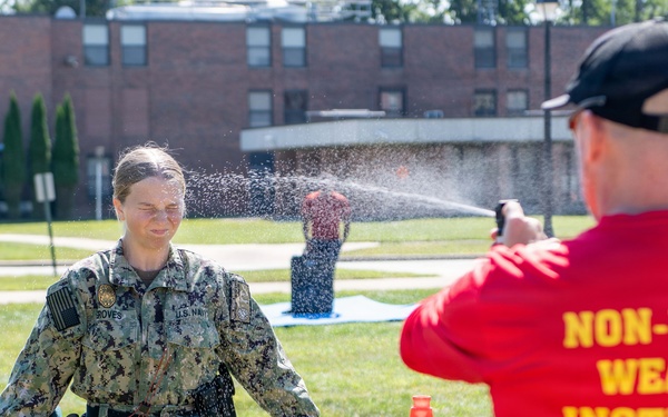 A U.S. Navy personnel member is being sprayed with what appears to be OC (pepper) spray or a similar non-lethal substance as part of a training exercise. A Non-Lethal Weapons Instructor in a red shirt is administering the spray. The training is taking place outdoors on a grassy area of what appears to be a military installation.