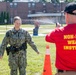 A U.S. Navy personnel member is being sprayed with what appears to be OC (pepper) spray or a similar non-lethal substance as part of a training exercise. A Non-Lethal Weapons Instructor in a red shirt is administering the spray. The training is taking place outdoors on a grassy area of what appears to be a military installation.