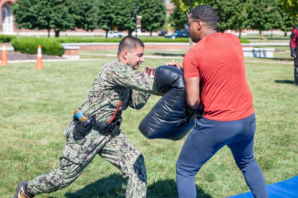 A person in U.S. Navy digital camouflage uniform with duty belt equipment is performing a combat or self-defense training drill outdoors on a grassy area. They are pushing against a large padded shield held by a civilian in a red shirt and blue pants. The setting appears to be a military installation or campus, with brick buildings and orange traffic cones visible in the background.