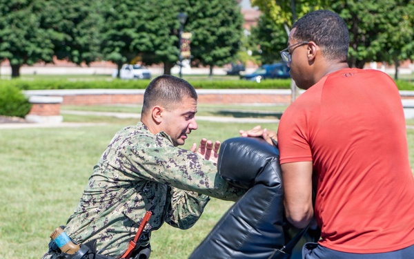 A person in U.S. Navy digital camouflage uniform with duty belt equipment is performing a combat or self-defense training drill outdoors on a grassy area. They are pushing against a large padded shield held by a civilian in a red shirt and blue pants. The setting appears to be a military installation or campus, with brick buildings and orange traffic cones visible in the background.