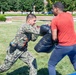 A person in U.S. Navy digital camouflage uniform with duty belt equipment is performing a combat or self-defense training drill outdoors on a grassy area. They are pushing against a large padded shield held by a civilian in a red shirt and blue pants. The setting appears to be a military installation or campus, with brick buildings and orange traffic cones visible in the background.