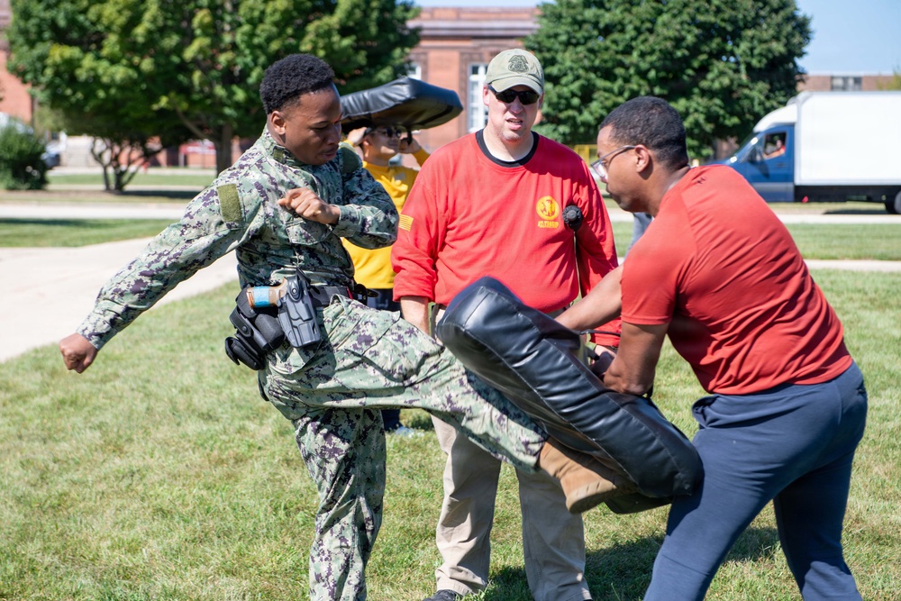 A military training exercise is taking place outdoors on a grassy area, likely on a military base. A person in Navy digital camouflage (NWU Type I) uniform is performing a kick against a padded training shield held by an instructor in a red shirt. A supervising instructor wearing a red shirt and green cap observes the technique.