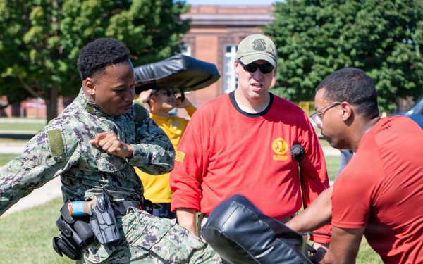 A military training exercise is taking place outdoors on a grassy area, likely on a military base. A person in Navy digital camouflage (NWU Type I) uniform is performing a kick against a padded training shield held by an instructor in a red shirt. A supervising instructor wearing a red shirt and green cap observes the technique.