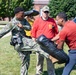A military training exercise is taking place outdoors on a grassy area, likely on a military base. A person in Navy digital camouflage (NWU Type I) uniform is performing a kick against a padded training shield held by an instructor in a red shirt. A supervising instructor wearing a red shirt and green cap observes the technique.