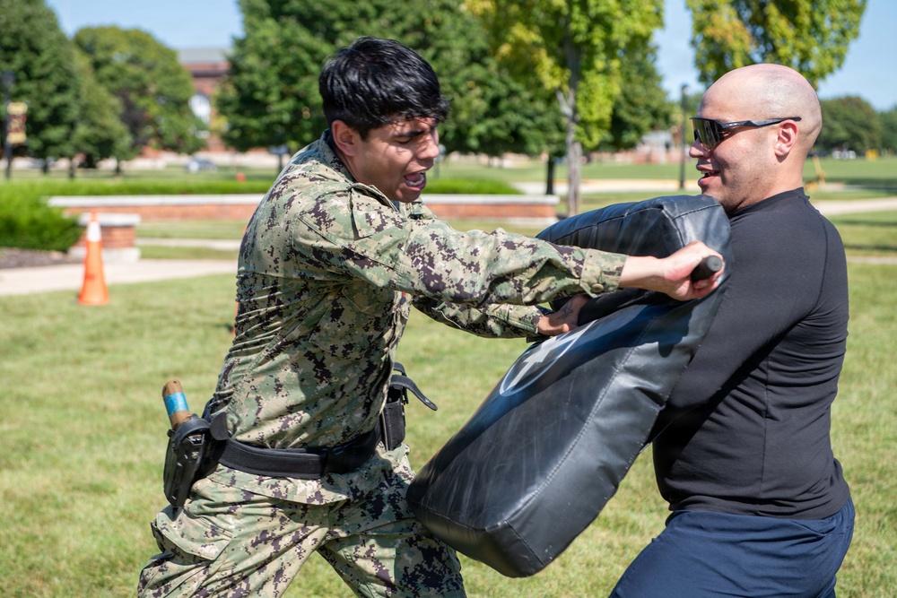 A military or law enforcement personnel in digital camouflage uniform is engaged in a combatives or defensive tactics training exercise outdoors. They are striking a padded training shield held by an instructor wearing a black long-sleeve shirt and sunglasses. The training appears to take place on a grassy area of what looks like a military base or institutional campus.
