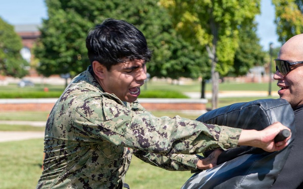 A military or law enforcement personnel in digital camouflage uniform is engaged in a combatives or defensive tactics training exercise outdoors. They are striking a padded training shield held by an instructor wearing a black long-sleeve shirt and sunglasses. The training appears to take place on a grassy area of what looks like a military base or institutional campus.