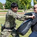 A military or law enforcement personnel in digital camouflage uniform is engaged in a combatives or defensive tactics training exercise outdoors. They are striking a padded training shield held by an instructor wearing a black long-sleeve shirt and sunglasses. The training appears to take place on a grassy area of what looks like a military base or institutional campus.