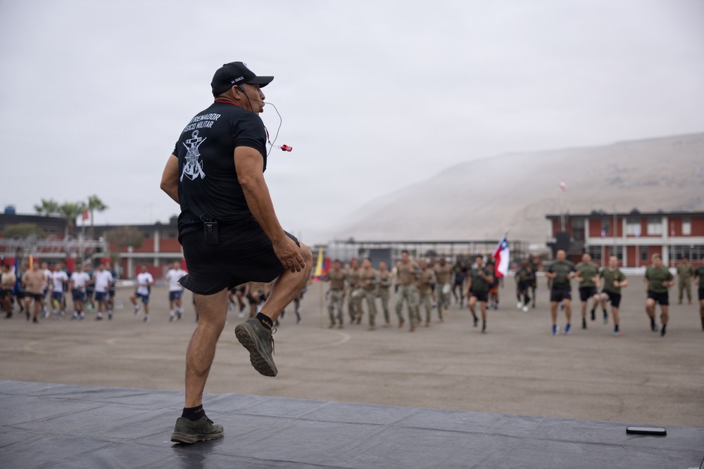 A military physical training instructor stands on an elevated platform leading a large group of soldiers in exercise drills on a parade ground. The instructor, wearing a black t-shirt with military insignia and a cap, is demonstrating a high-knee movement while holding a whistle. Multiple formations of military personnel in various uniforms are visible running or exercising in the background, with a Chilean flag visible among the group.