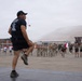 A military physical training instructor stands on an elevated platform leading a large group of soldiers in exercise drills on a parade ground. The instructor, wearing a black t-shirt with military insignia and a cap, is demonstrating a high-knee movement while holding a whistle. Multiple formations of military personnel in various uniforms are visible running or exercising in the background, with a Chilean flag visible among the group.