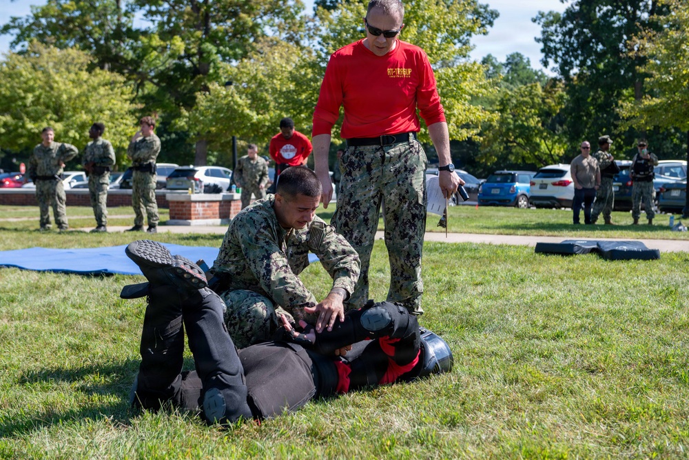 A military combatives or self-defense training exercise is taking place on a grassy outdoor area. A service member in Navy Working Uniform (NWU) camouflage is practicing a ground control technique on a person wearing protective padding and a helmet, while an instructor in a red shirt and camouflage pants observes and evaluates. Multiple other uniformed personnel stand in the background watching the demonstration.