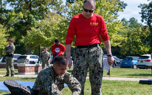 A military combatives or self-defense training exercise is taking place on a grassy outdoor area. A service member in Navy Working Uniform (NWU) camouflage is practicing a ground control technique on a person wearing protective padding and a helmet, while an instructor in a red shirt and camouflage pants observes and evaluates. Multiple other uniformed personnel stand in the background watching the demonstration.