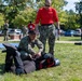 A military combatives or self-defense training exercise is taking place on a grassy outdoor area. A service member in Navy Working Uniform (NWU) camouflage is practicing a ground control technique on a person wearing protective padding and a helmet, while an instructor in a red shirt and camouflage pants observes and evaluates. Multiple other uniformed personnel stand in the background watching the demonstration.