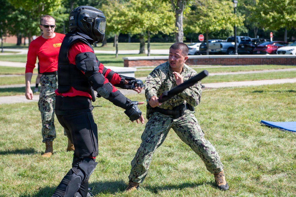A military training exercise is taking place outdoors on a grassy area. A sailor in Navy Working Uniform (NWU) camouflage is practicing baton/defensive tactics against a person wearing full protective padded training suit and helmet. A third person in a red shirt and camouflage pants observes and supervises the drill in the background.