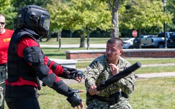 A military training exercise is taking place outdoors on a grassy area. A sailor in Navy Working Uniform (NWU) camouflage is practicing baton/defensive tactics against a person wearing full protective padded training suit and helmet. A third person in a red shirt and camouflage pants observes and supervises the drill in the background.