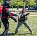 A military training exercise is taking place outdoors on a grassy area. A sailor in Navy Working Uniform (NWU) camouflage is practicing baton/defensive tactics against a person wearing full protective padded training suit and helmet. A third person in a red shirt and camouflage pants observes and supervises the drill in the background.