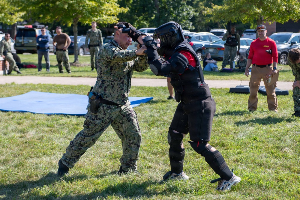 A military training exercise is taking place outdoors on a grassy field. A service member in Navy Working Uniform (NWU Type III camouflage) is engaged in hand-to-hand combat training with an instructor wearing full protective padding and a helmet. Multiple observers in military uniforms and civilian clothing watch from the background.