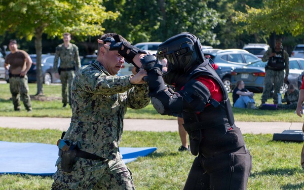 A military training exercise is taking place outdoors on a grassy field. A service member in Navy Working Uniform (NWU Type III camouflage) is engaged in hand-to-hand combat training with an instructor wearing full protective padding and a helmet. Multiple observers in military uniforms and civilian clothing watch from the background.