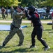 A military training exercise is taking place outdoors on a grassy field. A service member in Navy Working Uniform (NWU Type III camouflage) is engaged in hand-to-hand combat training with an instructor wearing full protective padding and a helmet. Multiple observers in military uniforms and civilian clothing watch from the background.