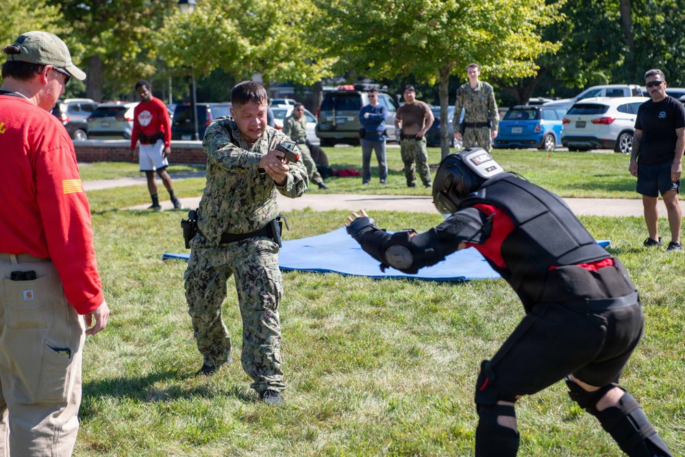 A military training exercise is taking place outdoors on a grassy area. A sailor in Navy Working Uniform (NWU) camouflage is pointing a training firearm at a person wearing full protective padded training gear (red and black suit with helmet). Several observers, including military personnel and civilians, watch from the background.