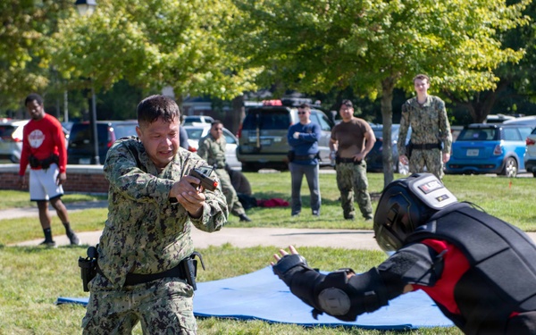 A military training exercise is taking place outdoors on a grassy area. A sailor in Navy Working Uniform (NWU) camouflage is pointing a training firearm at a person wearing full protective padded training gear (red and black suit with helmet). Several observers, including military personnel and civilians, watch from the background.
